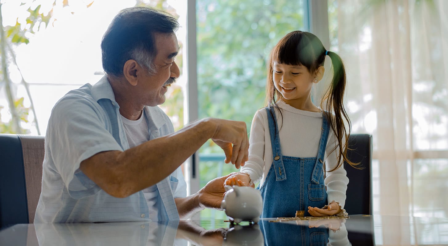 An older man and a little girl share a moment together at a table and giving her coins to teach the value of saving.