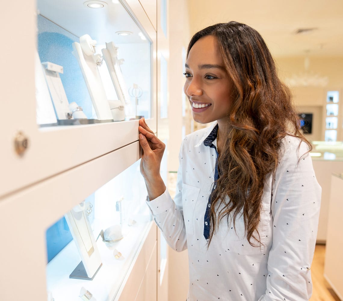 A woman wearing a white dress shirt examines various pieces of jewelry displayed in a store.