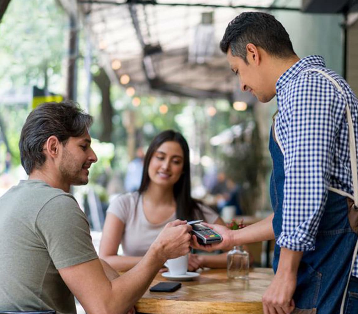 A man is making payment by tapping with his credit card as the waiter holds the mobile payment system in a restaurant setting. 