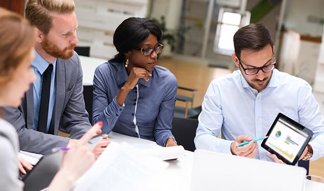 Four professionals are seated around a table in a meeting room. One man is holding a tablet, showing a graph to the group, while the others look on attentively. Papers and documents are on the table.