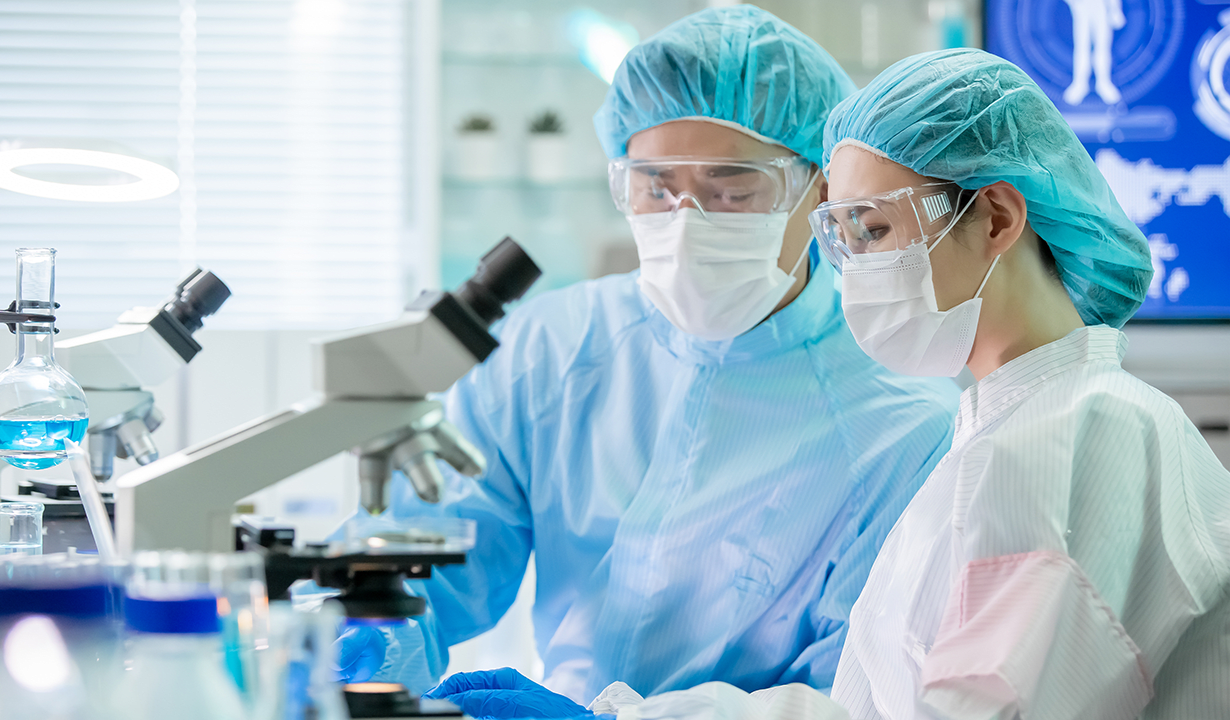 Two scientists in lab coats and masks examining samples under a microscope in a laboratory setting.
